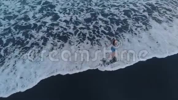 美丽的年轻女性在巴厘岛日落时带着黑色的沙滩和海浪沿着海滩散步飞机视频4K视频的预览图