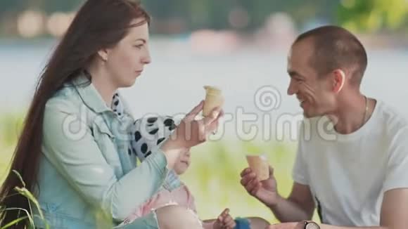 年轻快乐的父母带着一个小女儿坐在草地上玩着吃冰淇淋快乐视频的预览图