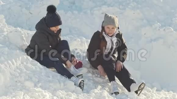 女孩们穿着溜冰鞋坐在溜冰场附近的雪堆里收紧鞋带视频的预览图