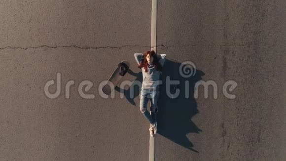 空中飞行一个带着滑板的少年躺在马路上用耳机听音乐年轻女孩嬉皮士视图视频的预览图