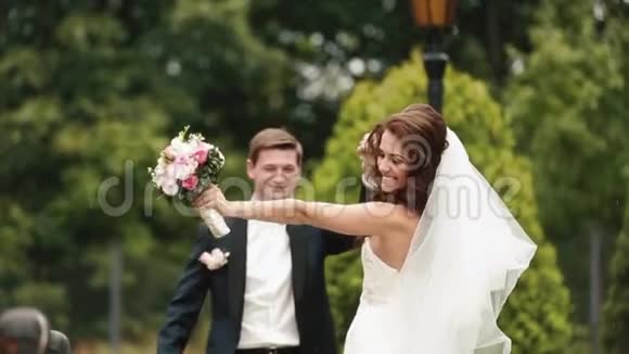美丽的一对新婚夫妇微笑着手牵着手走在雨中沿着绿色的绽放视频的预览图