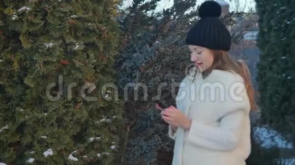 漂亮的金发碧眼穿着毛皮外套在FirTree后台的智能手机上发短信视频的预览图