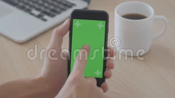 在工作场所的桌子咖啡和笔记本电脑上安装带有绿色屏幕Chroma键的男性手持智能手机视频的预览图