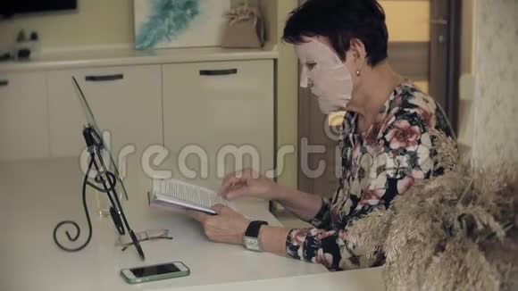 一位老妇人坐在桌子旁脸上戴着一个化妆织物面具她读一本书面部护理塑料视频的预览图