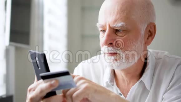 英俊的胡子老人坐在家里使用智能手机信用卡在线购物视频的预览图