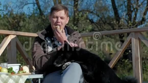 成熟的男人穿着温暖的衣服坐在桌子旁吃着汉堡靠近一只饥饿的狗在河岸上野餐视频的预览图