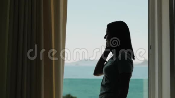 年轻女子用智能手机打电话的剪影高加索模特站在窗口拿着手机视频的预览图