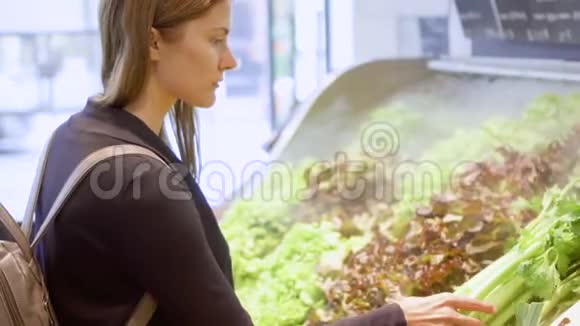 年轻女子在杂货店购物站在蔬菜部门选择新鲜的绿色芹菜视频的预览图