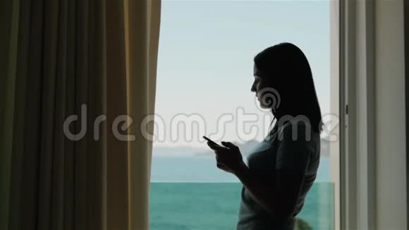 年轻女子智能手机剪影高加索模特站在窗口用她的智能手机视频的预览图