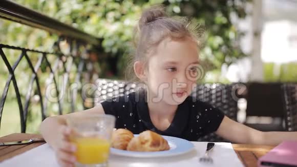 可爱的女孩在户外咖啡馆吃早餐可爱的女孩在露台上喝新鲜果汁和吃牛角面包视频的预览图