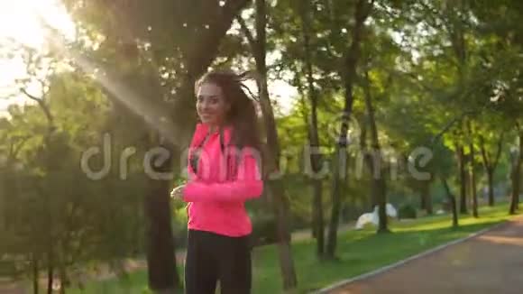 年轻的女人长发穿着明亮的粉红色夹克跑在阳光明媚的城市公园里看着镜头Stadicam视频的预览图