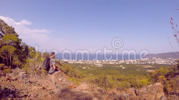 年轻人坐在山上望着城市徒步旅行者喝水观看美丽的大自然慢动作视频的预览图