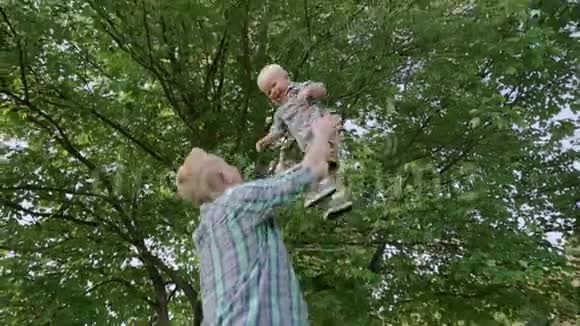 年轻的父亲牵着他的孩子的手在一个阳光明媚的日子里在公园里慢动作地把他扔到草地上视频的预览图