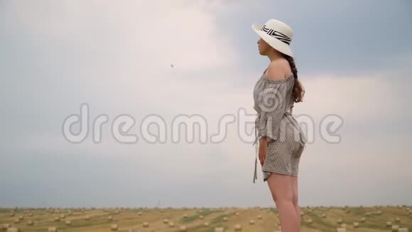 戴着帽子的年轻女子看着一片麦地的天空夏天的傍晚带着包视频的预览图