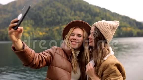两个漂亮的女孩穿着时髦的衣服在山上的湖边度假在手机上自拍视频的预览图