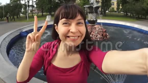 一位年轻女子在公园喷泉旁展示胜利标志自拍视频的预览图