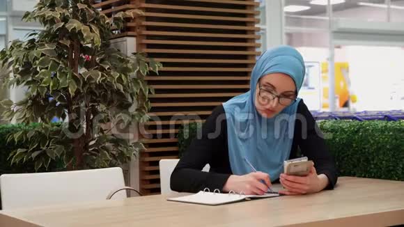 一个穆斯林女孩坐在咖啡馆里拿着手机在笔记本上写字视频的预览图