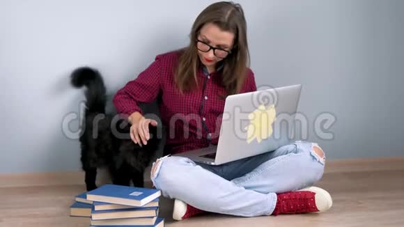 漂亮的女孩在家里坐在地板上用笔记本电脑抚摸一只黑猫视频的预览图