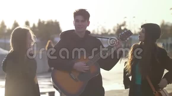 两个女孩微笑着而男人在户外日落时弹吉他视频的预览图