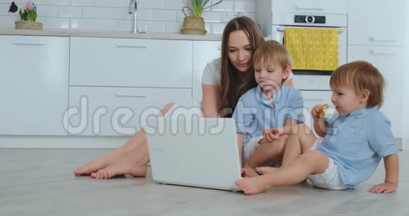 现代公寓妈妈和两个儿子坐在客厅的地板上看着笔记本电脑屏幕视频的预览图