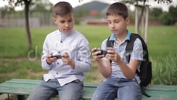 两个男孩坐在长凳上玩网络游戏一个带背包的男孩年轻人用电话视频的预览图