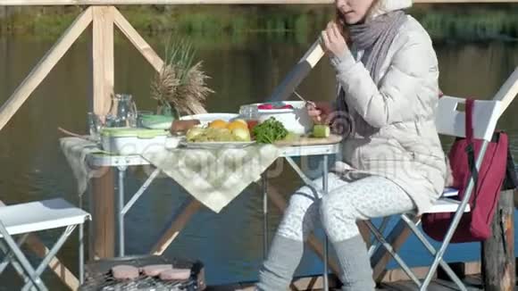 穿暖和衣服的年轻女子在河边的木桥上野餐切蔬菜周末寒冷的天气视频的预览图