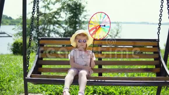 一个戴太阳镜的小可爱女孩在阳光明媚的温暖的日子里在跷跷板上摇摆视频的预览图