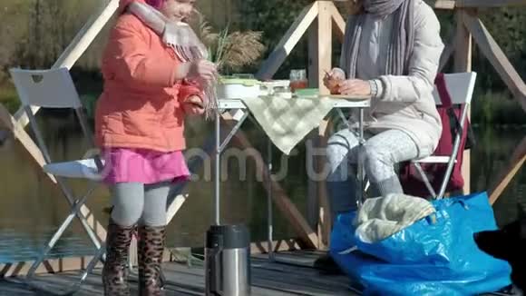 一个年轻的女人和一个穿暖和衣服的小女孩妈妈和女儿在一座木桥上的河岸上野餐视频的预览图