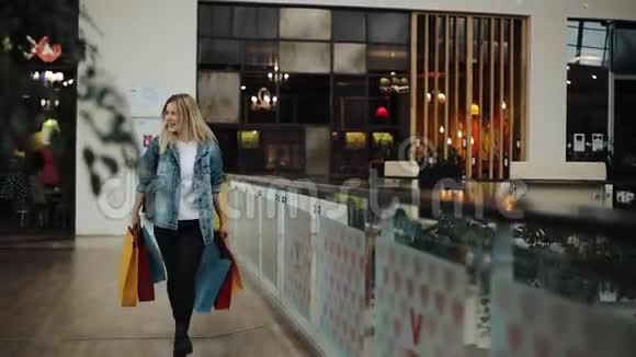 穿着牛仔裤夹克的金发女郎带着五颜六色的袋子在购物中心走来走去视频的预览图