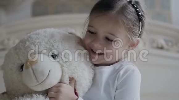 特写镜头小女孩坐在床上的白色地毯上抱着一个柔软的熊玩具漂亮的女孩和她的白色软玩具视频的预览图