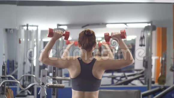 年轻漂亮的运动女孩用哑铃做肩部按压在运动健身房锻炼后侧视视频的预览图