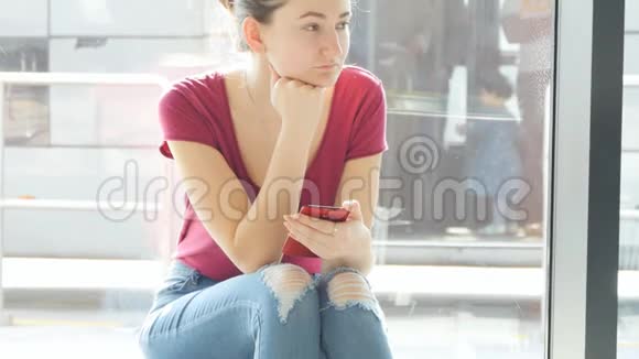 年轻女子坐在机场候机室的窗户附近环顾四周然后使用智能手机预期视频的预览图