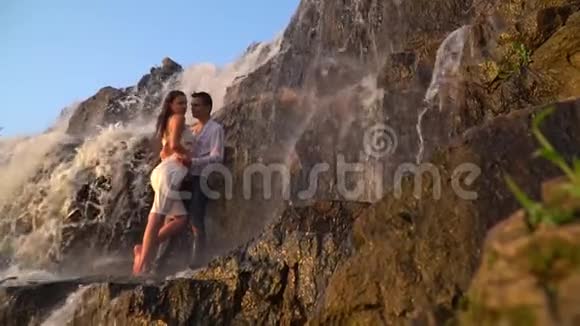 一个夏天的日落时分一个穿着湿衣服的男人和一个女孩站在瀑布下的岩石中动作缓慢视频的预览图