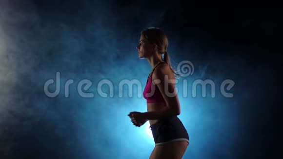 从侧面看奔跑的女孩在烟雾弥漫的黑色聚光灯背景慢动作视频的预览图