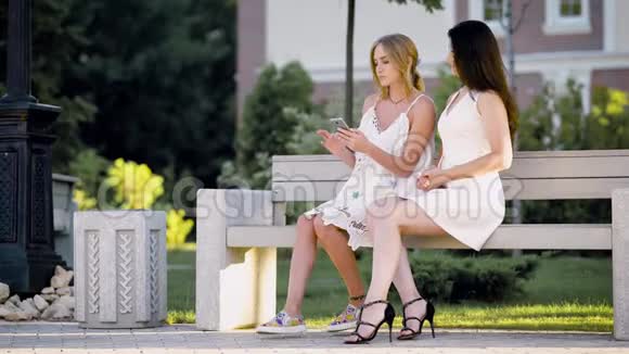 年轻时尚的白人女性在街上的长凳上快乐地摆姿势一起浏览智能手机视频的预览图