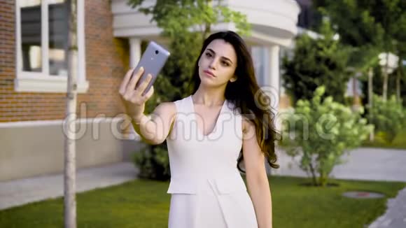 年轻严肃的黑发穿着白色连衣裙在街头背景下摆出自拍的智能手机视频的预览图