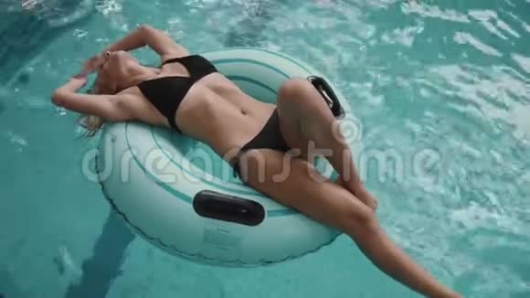 迷人的女人穿着黑色比基尼在游泳池里的充气橡胶圈上游泳4k视频的预览图