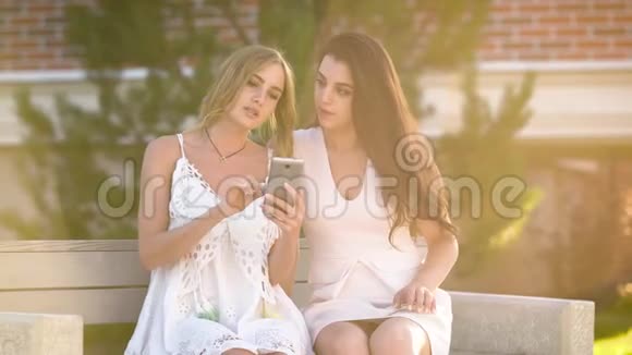 两个穿着白色连衣裙的时髦女孩坐在长凳上在夏天的阳光下分享智能手机视频的预览图