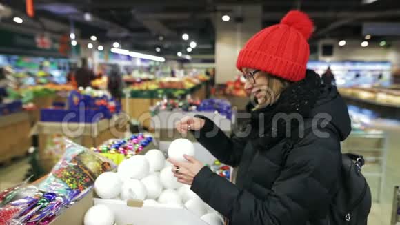 年轻漂亮的白种人女孩戴着眼镜戴着红帽子在市场商店里挑选新年树的球视频的预览图