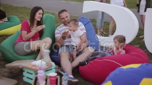 快乐的年轻家庭有两个孩子在公园野餐男孩和女孩吃披萨坐在豆袋椅子上视频的预览图