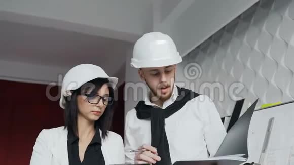 两名年轻建筑师他们在建筑工地上戴着白盔讨论他们在建筑工地上提供的信息视频的预览图