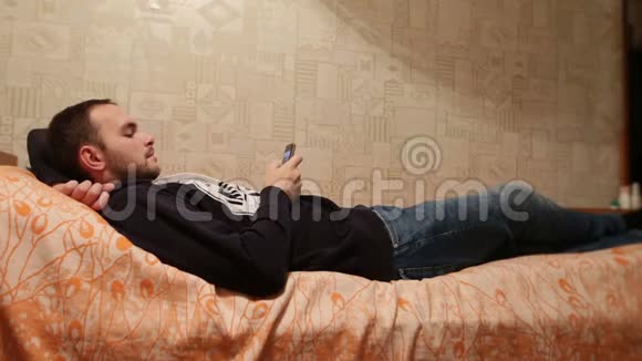 男人躺在家里的床上在肖像模式下使用手机懒惰的白种人在看新闻或照片视频的预览图