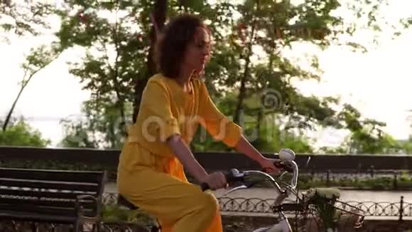 穿着黄色长裙的漂亮女人在夏天骑自行车穿过城市车上有篮子和鲜花视频的预览图