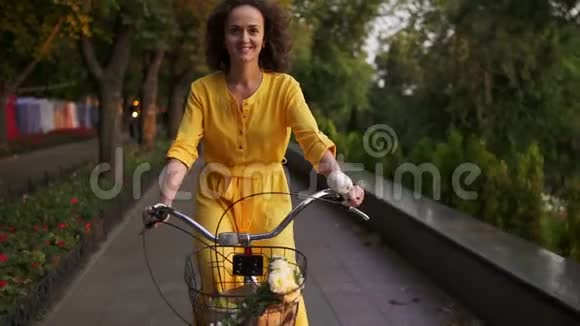 美丽的女人穿着黄色的长裙骑着一辆城市自行车篮子和鲜花在城市公园里看着视频的预览图