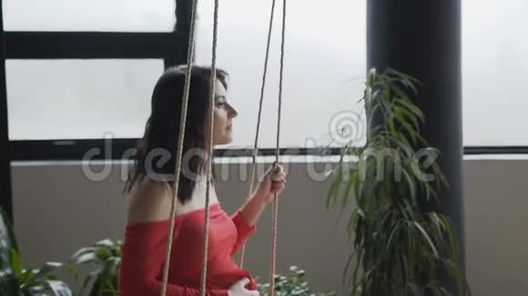从孕妇的视野她穿着一件红色的长裙在大窗户附近的秋千上摆动怀孕概念视频的预览图