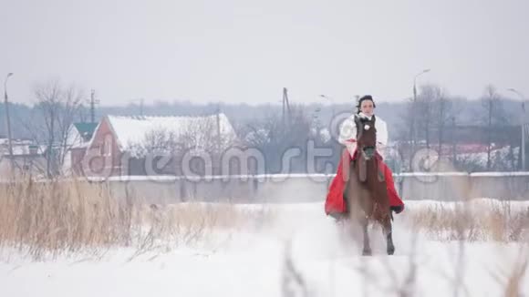 穿着红色连衣裙的漂亮黑发年轻姑娘在冬天的雪地里飞快地骑在马背上视频的预览图