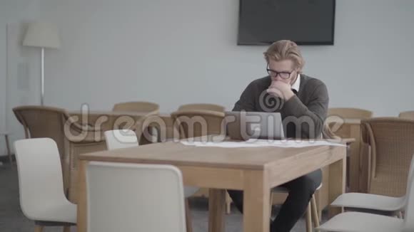 金发体贴的男人戴着眼镜坐在上网本前一间舒适的办公室里的桌子旁帅哥视频的预览图