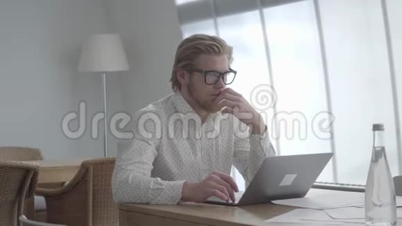 金发体贴的男人戴着眼镜坐在上网本前一间舒适的办公室里的桌子旁帅哥视频的预览图