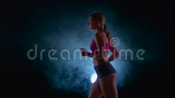 女孩跑在黑色的背景上被烟雾中的聚光灯照亮慢动作关门视频的预览图