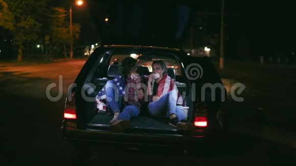 美丽的女孩坐在敞开的汽车后备箱里在城市的夜晚聊天和吃汉堡视频的预览图